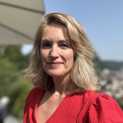 Frau mit schulterlangen blonden Haaren und rotem T-Shirt blickt freundlich in die Kamera, im Hintergrund verschwommen ein Sonnenschirm.