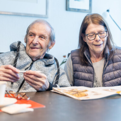 Mann mit grauen Haaren und grauer Kapuzenjacke hält Stift in der Hand, Frau mit braunen Haaren und gesteppter Weste sitzt daneben am Tisch mit aufgeschlagener Zeitschrift und Kaffeetasse