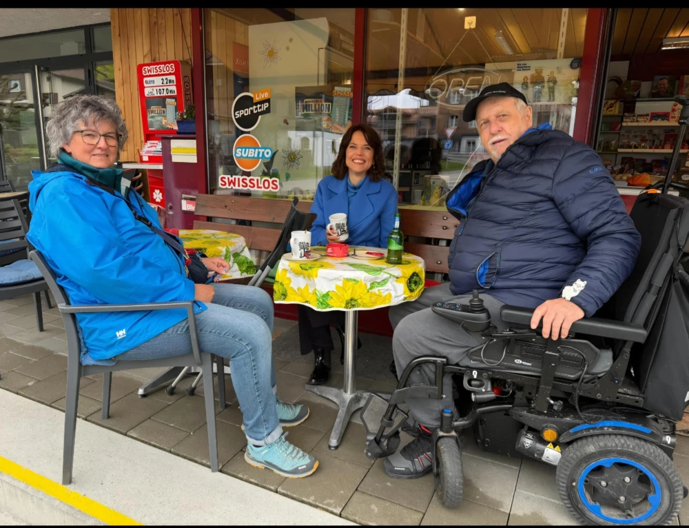 Annelise Engetschwiler, Mona Vetsch und Ruedi Däpp sitzen an einem runden Tisch mit gelb-grün gemusterter Tischdecke vor einem Café, Ruedi Däpp sitzt im Elektrorollstuhl.
