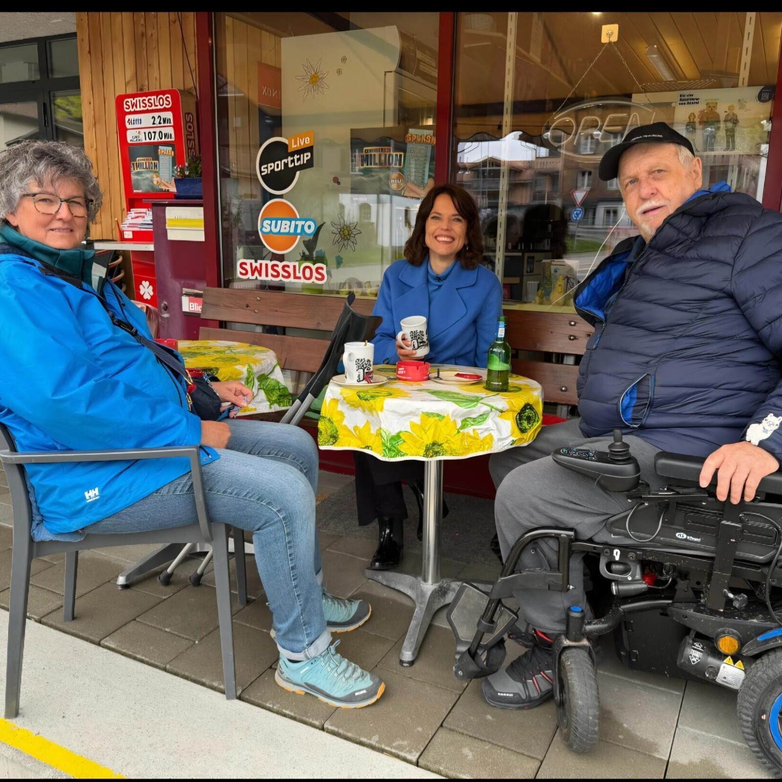 Annelise Engetschwiler, Mona Vetsch und Ruedi Däpp sitzen an einem runden Tisch mit gelb-grün gemusterter Tischdecke vor einem Café, Ruedi Däpp sitzt im Elektrorollstuhl.