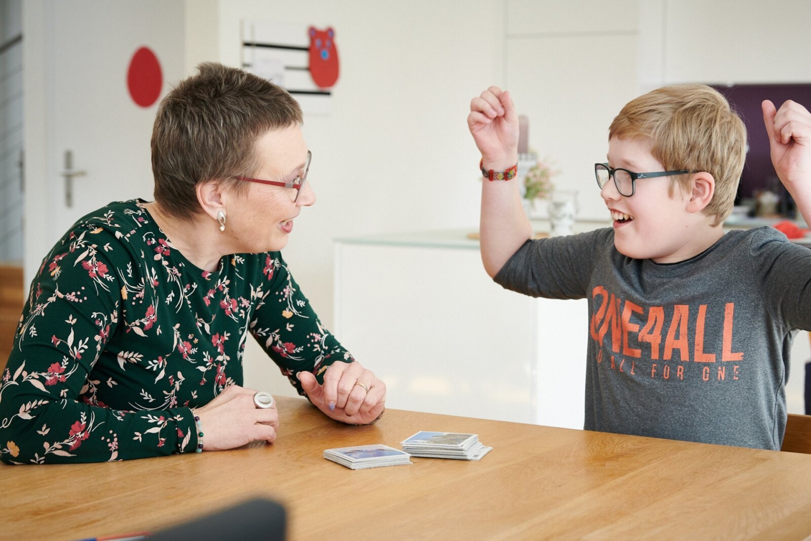 Eine Frau spielt ein Kartenspiel mit einem Jungen mit Beeinträchtigung, der seine Arme freudig in die Luft streckt